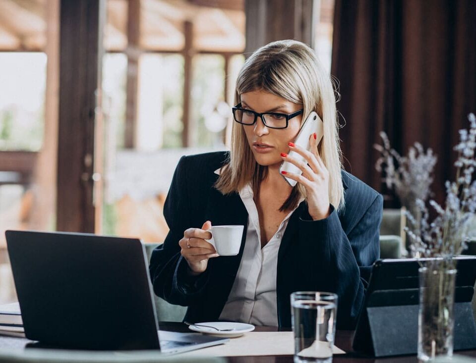 zena sedi ispred laptopa, prica na telefon i pije kafu