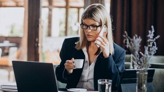 zena sedi ispred laptopa, prica na telefon i pije kafu