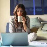 Zena u sivoj jakni sedi na krevetu, pije kafu i radi na laptopu