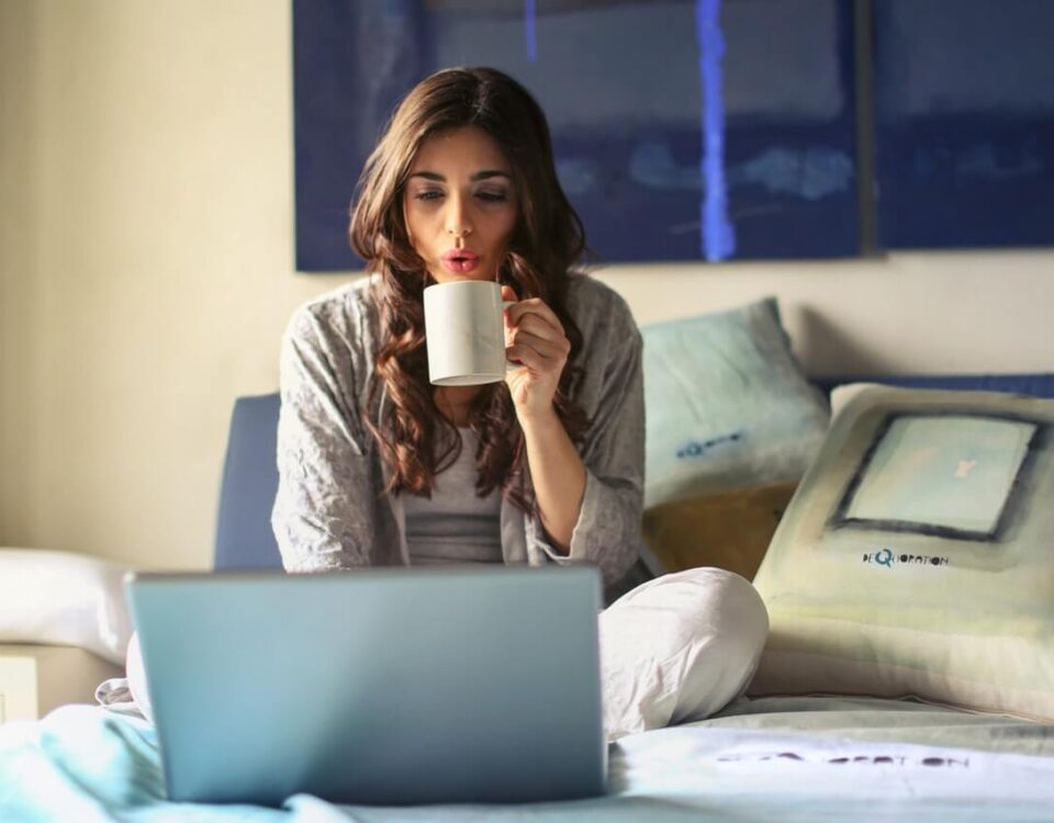 Zena u sivoj jakni sedi na krevetu, pije kafu i radi na laptopu